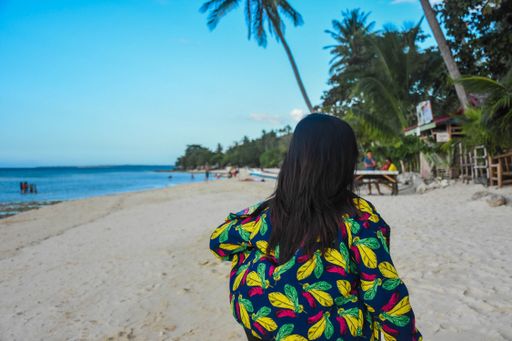 A female in a coat with colorful feathers walking on a populated beach with beautiful natural scenery in the background