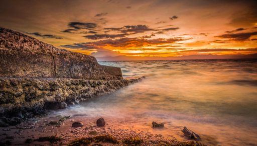 A beautiful wide shot of coast of the sea with rocks on the right and amazing breathtaking clouds in the red sky during sunset