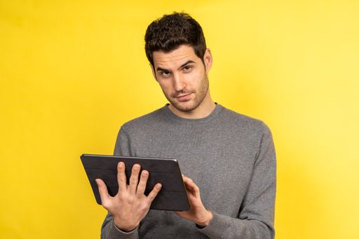 A young Caucasian male with a gray shirt working on the tablet and looking at the camera against an orange background