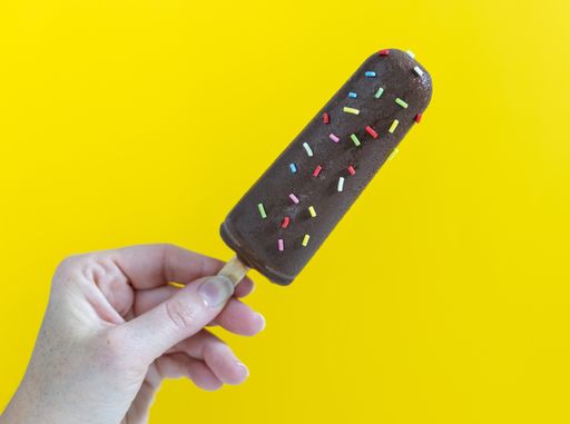 Woman's hand taking a chocolate ice cream covered with colored nuggets on a yellow background