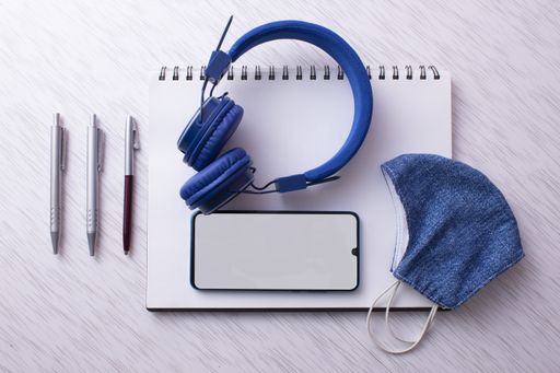 A top view of  a cell phone with headphones, notebook, and medical mask on a surface