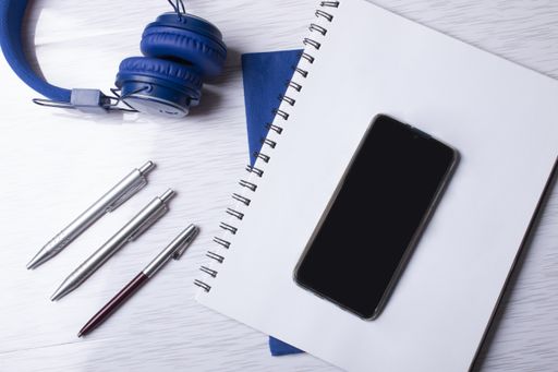 A top view of  a cell phone with headphones, notebooks, and pens on a surface