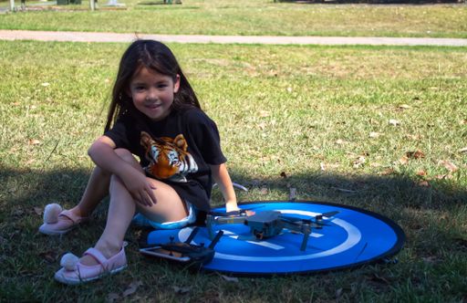 A closeup of a little girl sitting next to a drone in the shadows on a sunny day