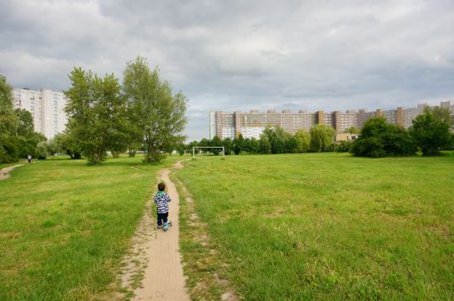 Young boy riding a scooter on a small footpath along green grass and apartment building in the distance.