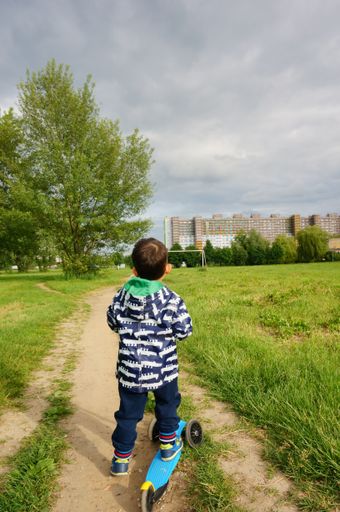 Young boy riding a scooter on a small footpath along green grass and apartment building in the distance.