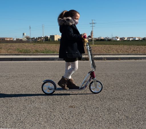 A cute Caucasian little girl in a face mask riding her scooter through the paved street