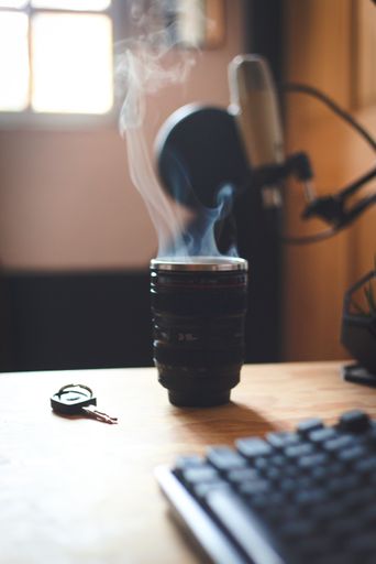 A vertical shot of hot coffee in a camera lens mug on the table with a microphone in the background