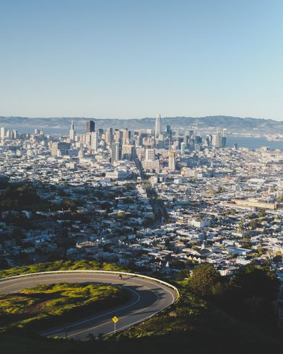 A vertical shot of a curvy road down the hill with city buildings in the distance under a blue sky