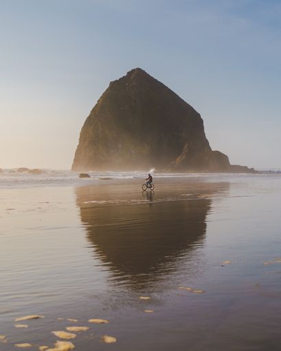 A vertical shot of a person riding a bicycle on the seashore with a big rock in the distance