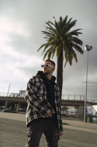 A vertical shot of a stylish guy wearing a plaid jacket, a black hoodie, black pants, and sunglasses