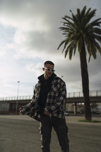 A vertical shot of a stylish guy wearing a plaid jacket, a black hoodie, black pants, and sunglasses