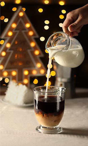 A vertical shot of a woman pouring milk into coffee with a Christmas-themed decorations in the background