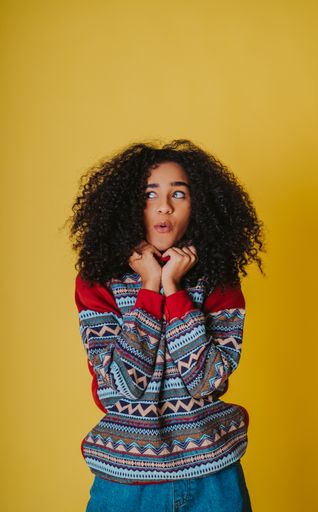 A cheerful Hispanic female with curly hair wearing jeans and a hoodie and posing against a yellow background