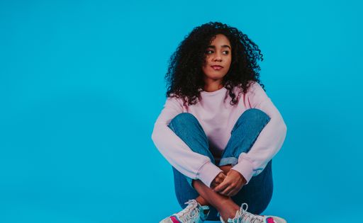 A young Hispanic female with curly hair wearing a hoodie with jeans and sitting against a blue background