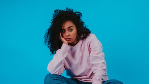 A thoughtful Hispanic female wearing a hoodie with jeans and sitting against a blue background