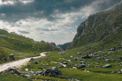 A gravel road surrounded by grass-covered lands and mountains on a cloudy day