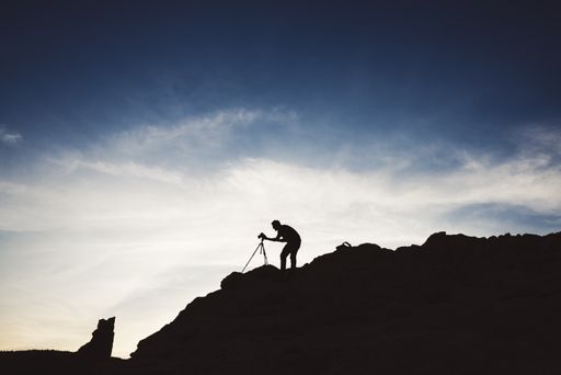 A silhouette of a photographer standing on a hill with a camera on a stand under the sunlight in the evening