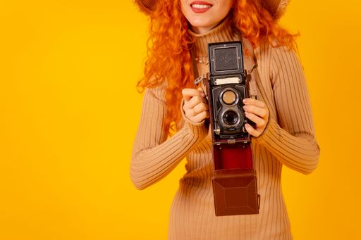 A closeup of a red-haired Caucasian tourist with a hat and vintage camera on a yellow background, with copy space