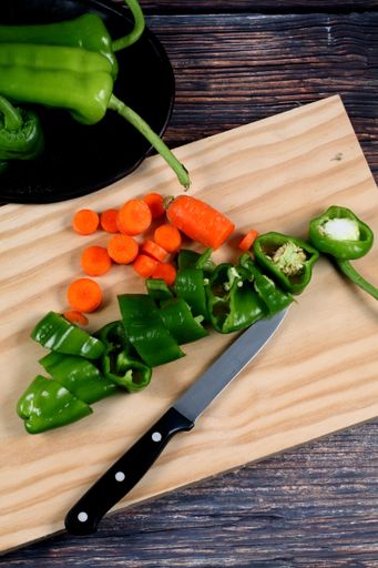 A vertical shot of a knife, chopped carrots peppers on a wooden board, and green peppers on a plate