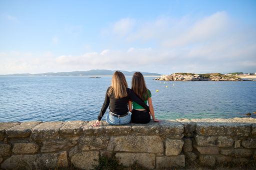 A mother and a daughter on their backs sitting on a stone bench looking at the sea