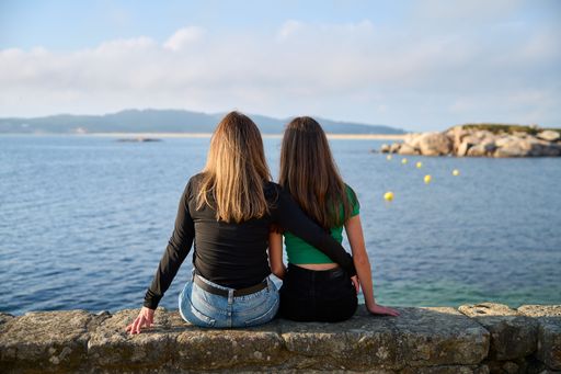 A mother and a daughter on their backs sitting on a stone bench looking at the sea