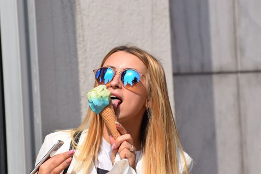 A young beautiful blonde girl with sunglasses licking a yellow and blue ice cream cone in the street