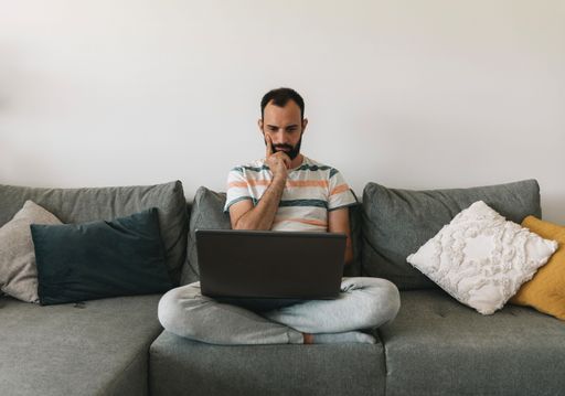 A middle-aged male with facial hair sitting at home on the couch and working on a laptop