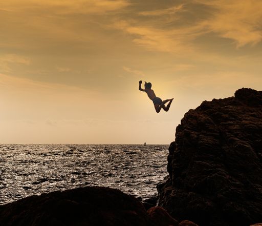 An excited young boy jumping from a rocky cliff into the lake water surrounded by a beautiful sunset view