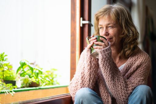 A Caucasian senior woman with blonde hair drinking tea and looking out from the window
