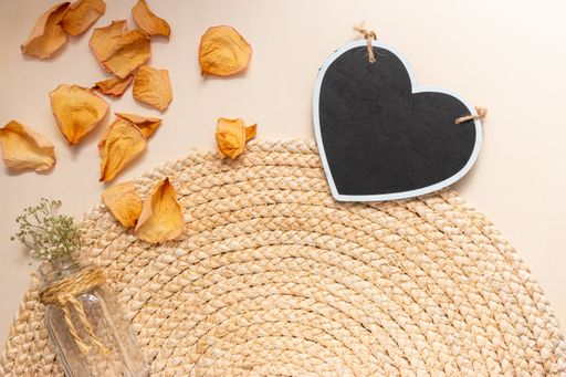 The flat lay of a glass vase with flowers, black heart-shaped board, and dry rose petals on the round jute table mat, on the beige backgrou