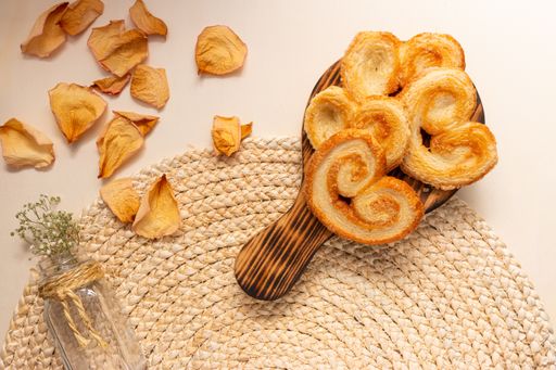 The flat lay of a glass vase with flowers, a board with palmier cookies, and dry rose petals on the round jute table mat, on the beige background