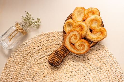 The flat lay of a glass vase with flowers and a board with palmier cookies on the round jute table mat, dessert for St. Valentine's day