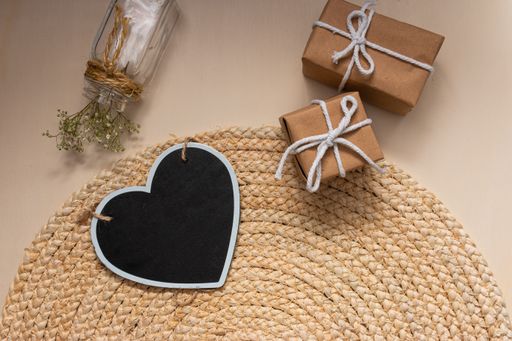The flat lay of a black heart-shaped board and gift boxes on the round jute table mat, a glass vase with flowers on the beige background