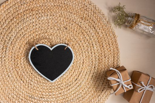 The flat lay of a black heart-shaped board on the round jute table mat, gift boxes and a glass vase with flowers on the beige background