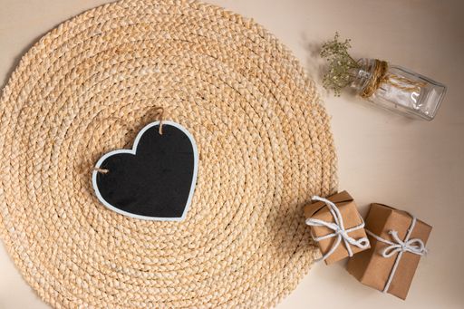 A composition with a round jute table mat and a black heart-shaped board, a glass vase with flowers and gift boxes on the beige background