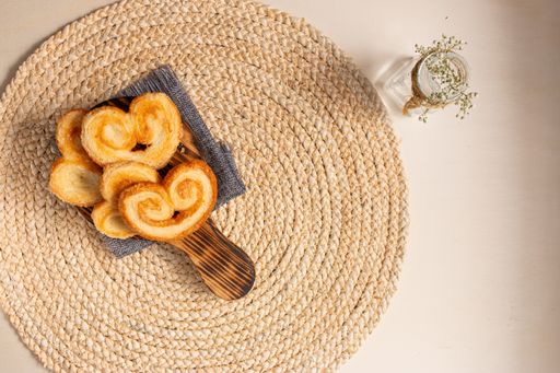 The flat lay of yummy palmier cookies on the round jute table mat, flowers in a vase on the beige background