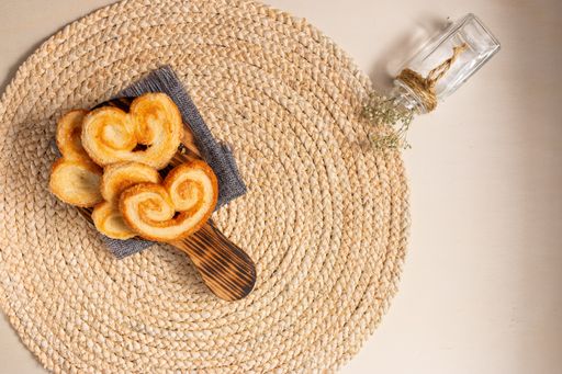 The flat lay of yummy palmier cookies on the round jute table mat, a small decorative flower vase on the right side of the beige background