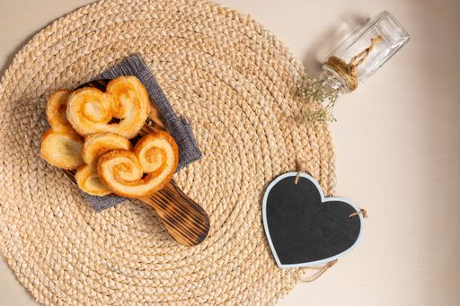 The flat lay of yummy palmier cookies and a black heart-shaped board on the round jute table mat, flowers in a vase on the beige background