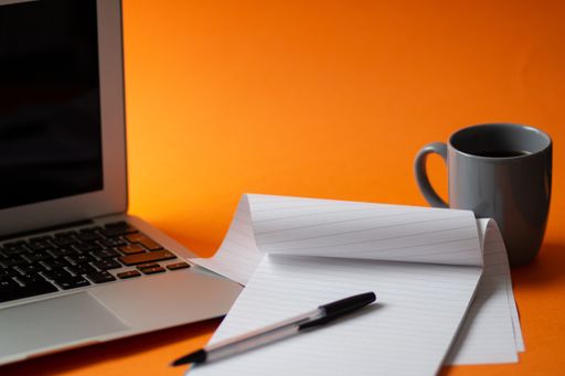 A selective focus shot of a laptop, notepad, pen and a cup of hot coffee isolated on an orange background - work from home concept