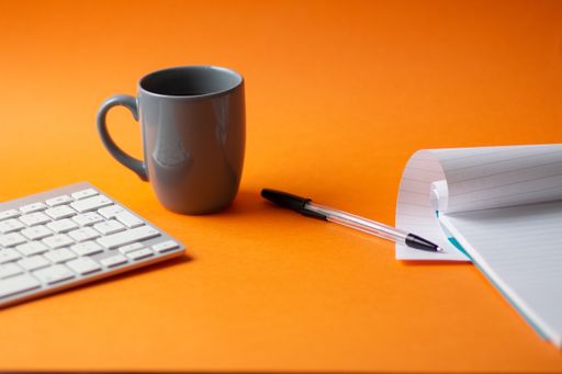 A selective focus shot of a keyboard, notepad, pen and a cup of hot coffee isolated on an orange background - work from home concept