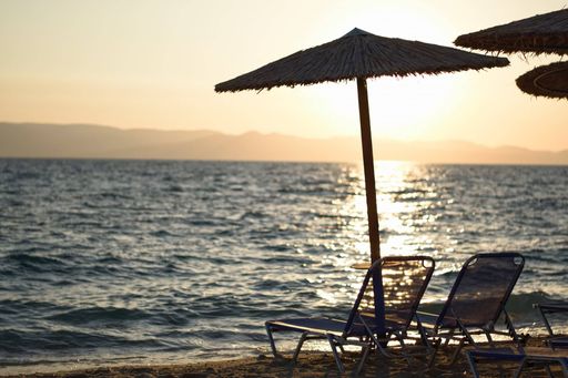 A selective focus of beach chairs under straw umbrellas with a sunset view over the blue sea in the backdrop