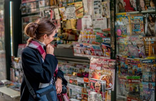 A thoughtful woman looking at the magazine showcase of a street kiosk with interest.