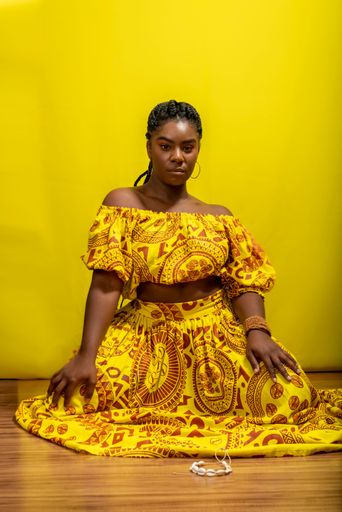 Young beautiful woman, serious, looking at the camera, sitting on a wooden floor. Against yellow background.