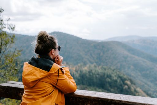 A female adult wearing a bright yellow jacket and sunglasses on a wooden balcony with a view of lush green trees in the background