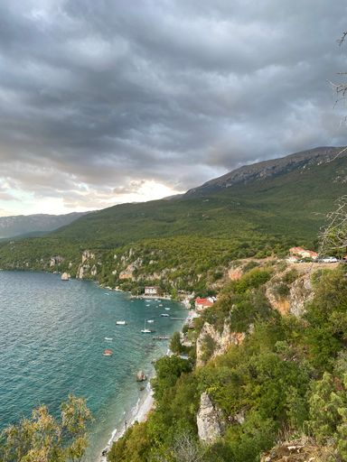 A vertical aerial shot of a coastline with cliffs and hill forests overlooking the sea, and yachts near the beach on a cloudy day
