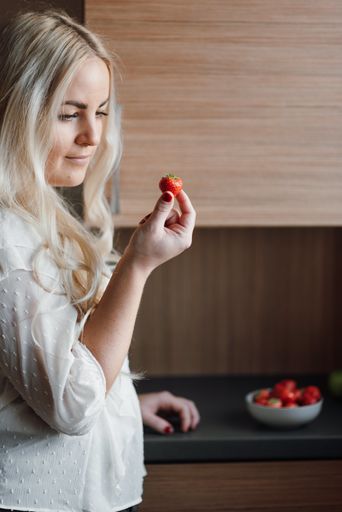A shallow focus of a blonde woman holding a strawberry in the kitchen
