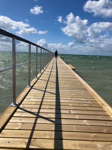 A man on a weathered pier that stretches out to the sea, with a cloudy sky serving as the backdrop