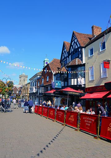 People walking and sitting at outdoor cafe tables, in the summer sunshine in the High Street, Salisbury, United Kingdom. Portrait/upright format image