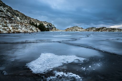 A sea covered in ice surrounded by rocks and houses under a cloudy sky during winter