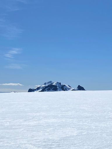 A majestic landscape featuring a large expanse of snow-covered terrain and two distant mountain peaks set against a backdrop of clear blue skies